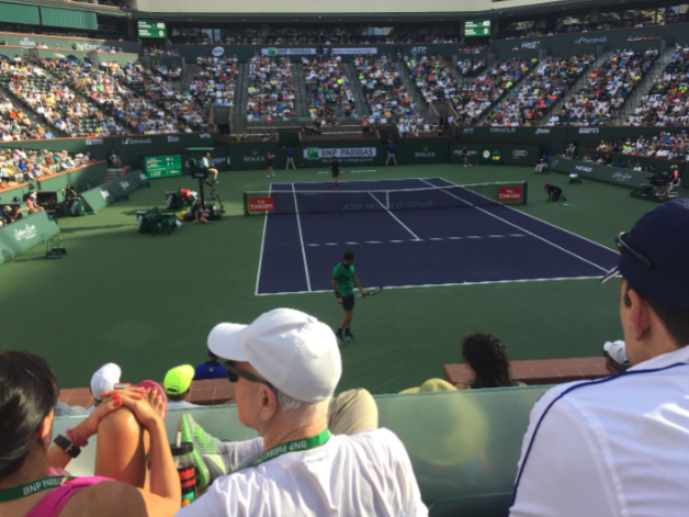 Tennis stadium with a green court and a player ready to serve, seen from the audience seats.